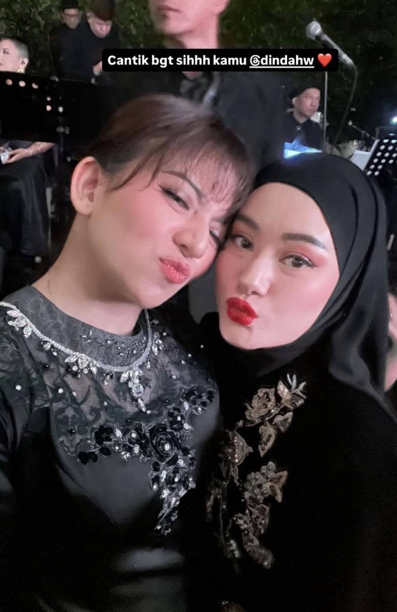 Dua wanita berpose bersama mengenakan busana hitam berhias payet di acara pernikahan dengan latar suasana malam dan musisi di belakang.