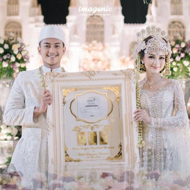 Pasangan pengantin mengenakan busana adat Sunda berwarna putih sambil memegang bingkai besar bersertifikat di acara pernikahan mewah.