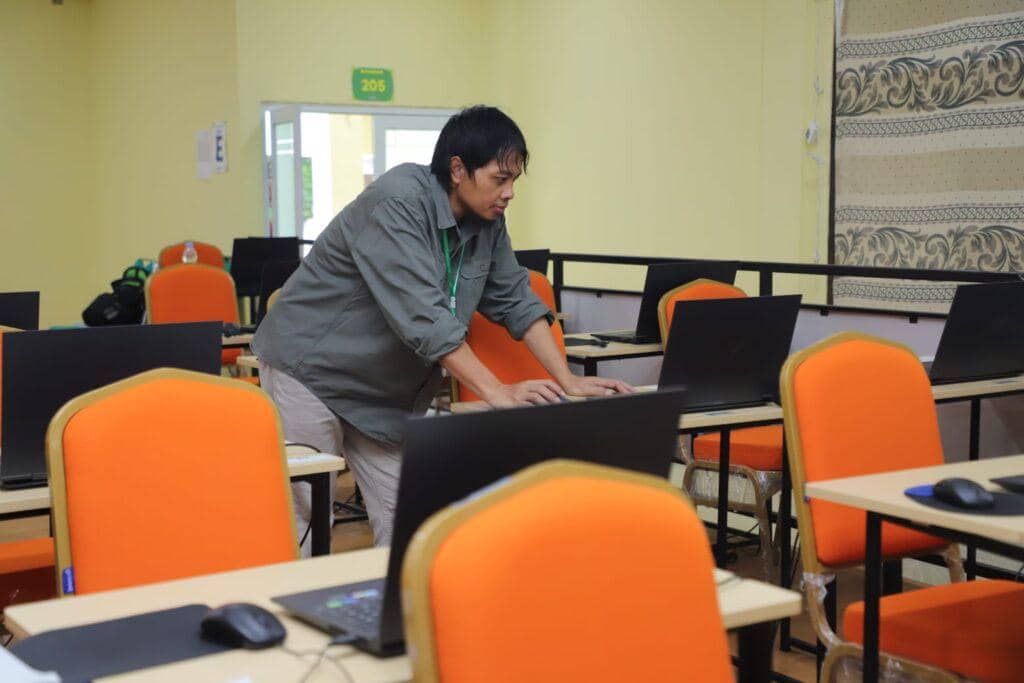 Seorang petugas memeriksa komputer di ruang ujian dengan deretan kursi oranye di Universitas Lampung saat pelaksanaan UTBK SNBT.