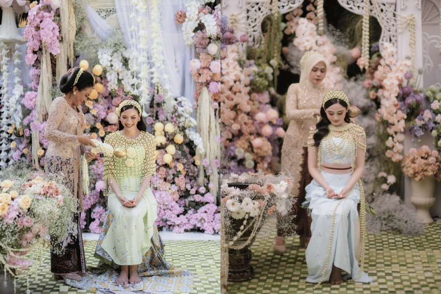 Dua perempuan mengenakan busana adat Jawa dalam prosesi siraman pernikahan, dikelilingi dekorasi bunga berwarna pastel yang indah.