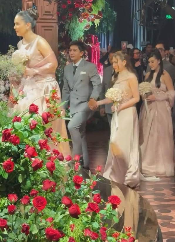 Seorang pria mengenakan setelan jas abu-abu berjalan bergandengan tangan dengan wanita bergaun krem di acara pernikahan bertema bunga mawar merah.