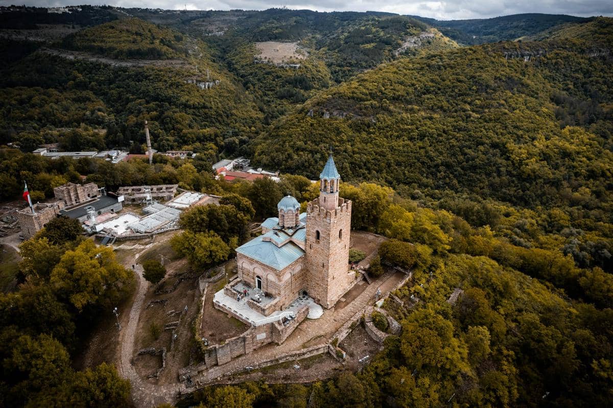 Pemandangan udara Veliko Tarnovo dengan bangunan bersejarah berdinding batu di tengah perbukitan hijau yang rimbun.