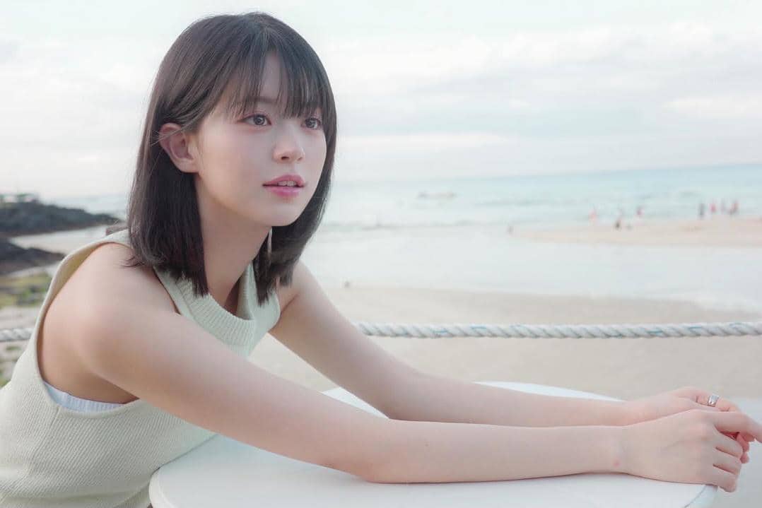Seorang perempuan berambut pendek mengenakan atasan tanpa lengan berwarna terang duduk di tepi meja dengan latar pantai yang tenang.
