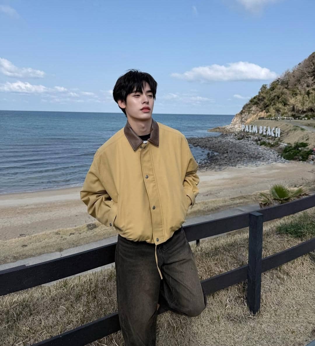 Seorang pria mengenakan jaket cokelat muda dan celana gelap berpose di tepi pantai dengan latar laut biru dan tulisan Palm Beach di bukit.
