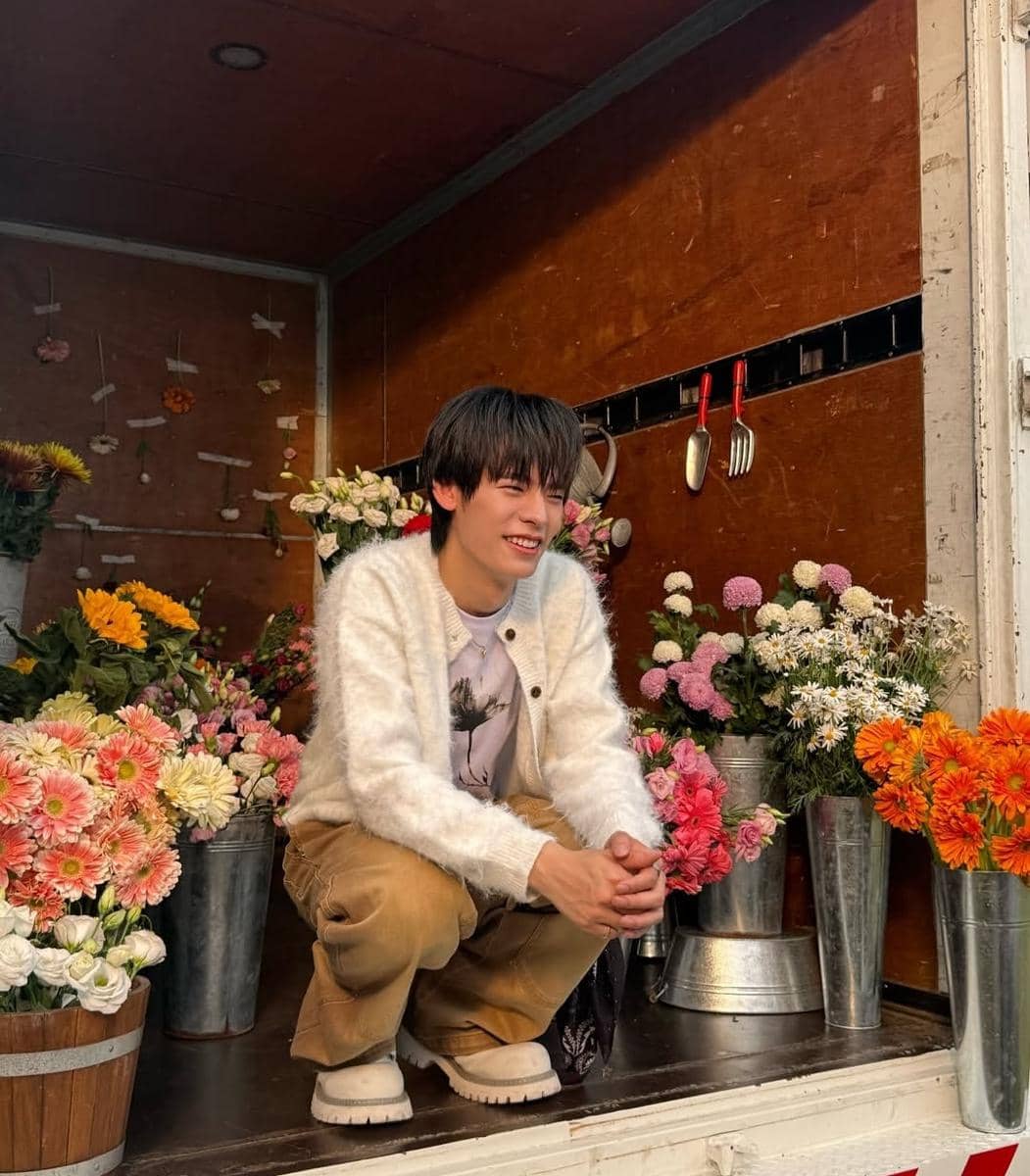 Seorang pria berpose santai di dalam truk penuh bunga warna-warni, mengenakan cardigan putih, kaus bergambar, dan celana cokelat.