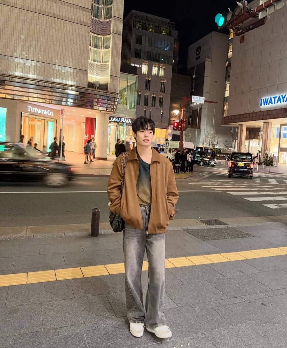 Seorang pria bergaya kasual mengenakan jaket cokelat dan celana jeans abu-abu berdiri di trotoar kota pada malam hari dengan latar gedung bertingkat.