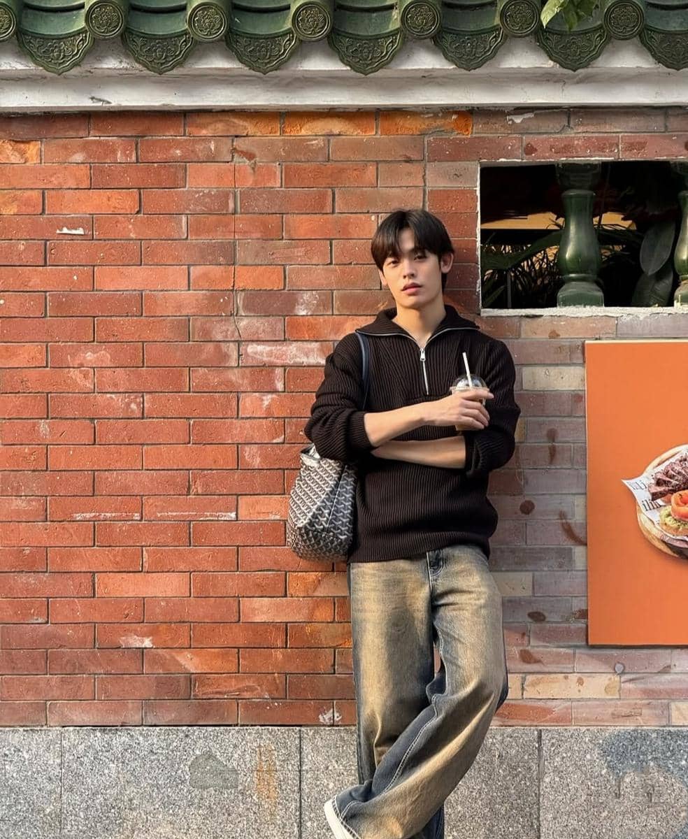 Seorang pria mengenakan sweater hitam dan celana jeans longgar sambil memegang minuman dan bersandar di dinding bata merah.