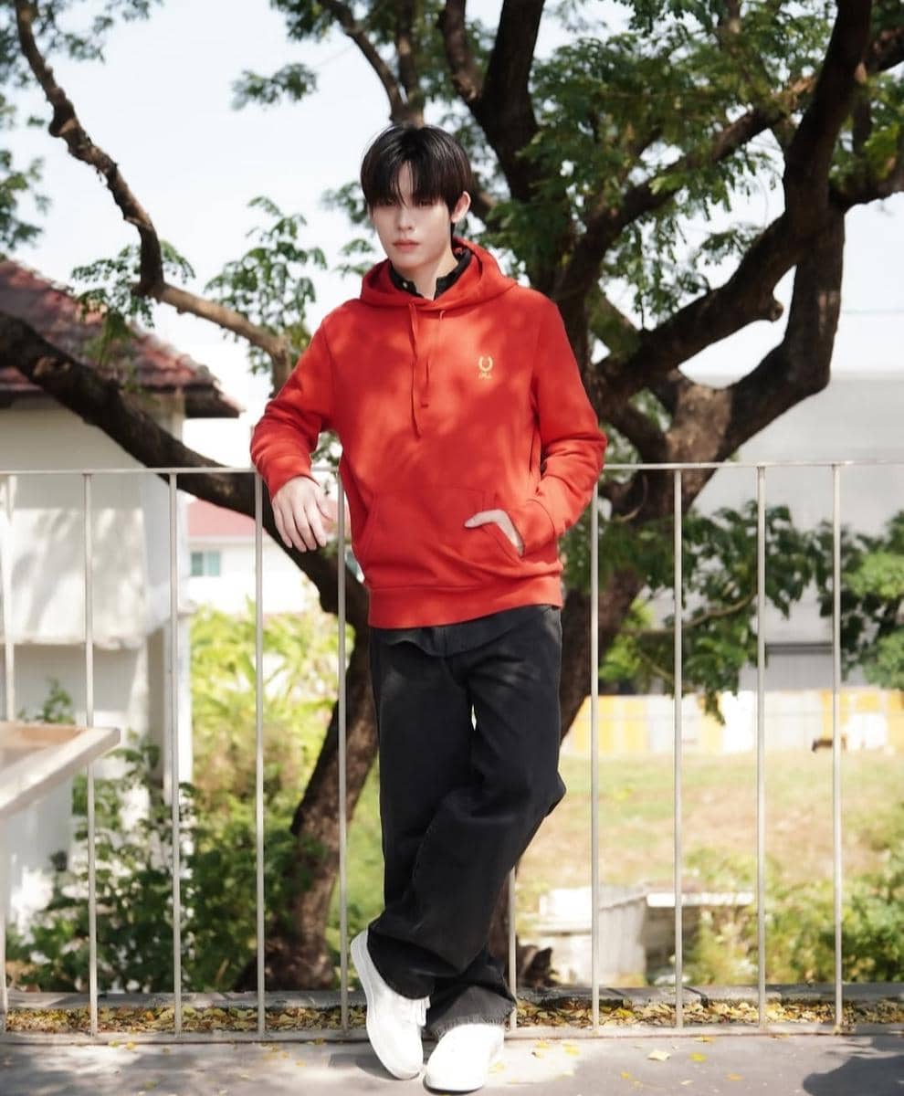 Seorang pria mengenakan hoodie oranye, celana panjang hitam, dan sepatu putih berdiri santai di luar ruangan dengan latar pepohonan.