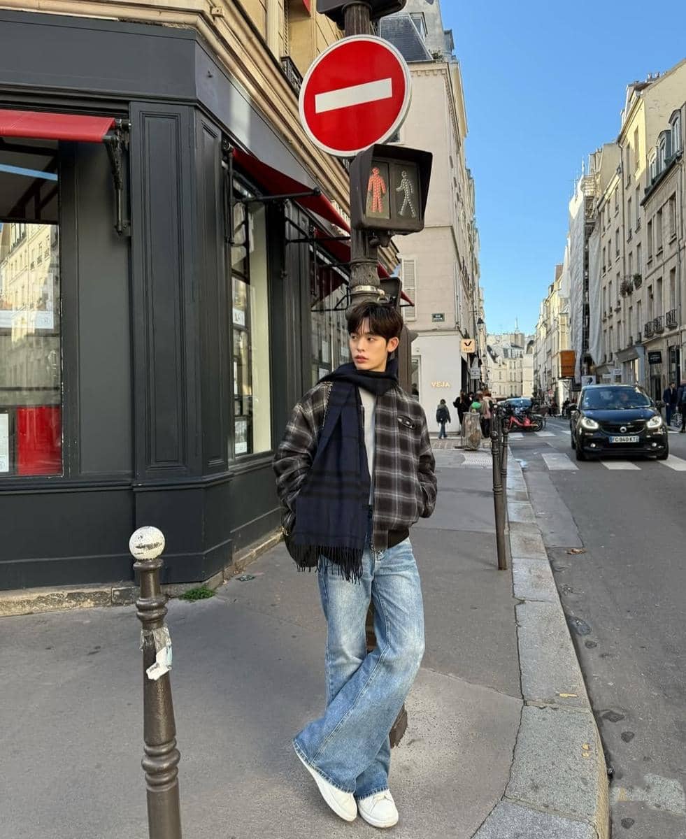 Seorang pria bergaya kasual dengan jaket kotak-kotak, syal gelap, dan jeans longgar berdiri di trotoar kota dengan latar bangunan Eropa.