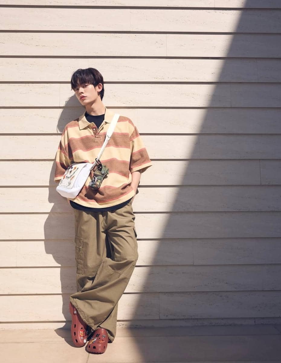 Seorang pria bergaya kasual mengenakan kaus garis-garis, celana kargo longgar, dan tas selempang putih sambil bersandar di dinding.