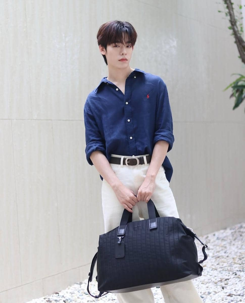 Pria mengenakan kemeja biru tua dan celana panjang putih sambil memegang tas besar hitam di area luar ruangan berdinding terang.