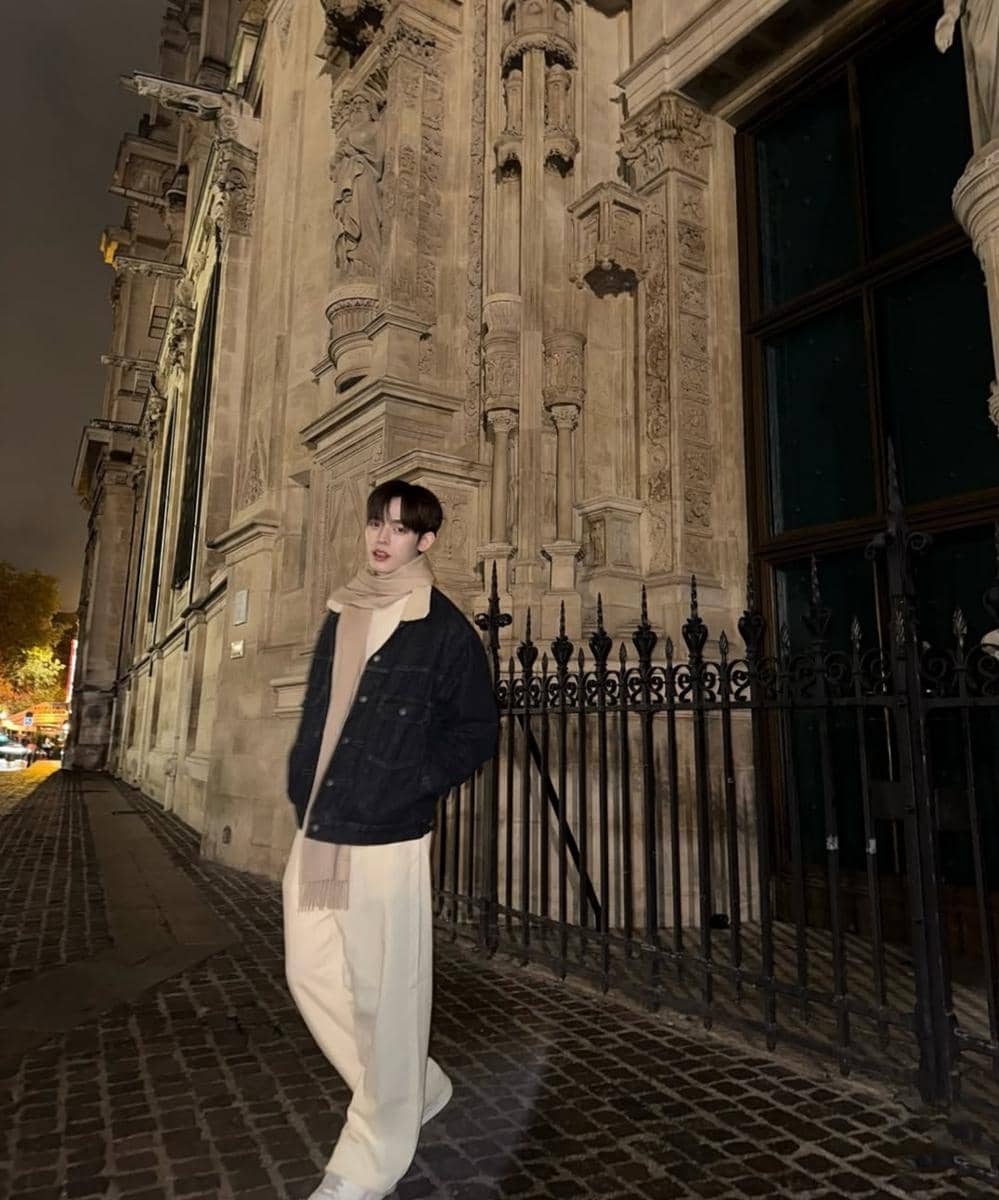 Seorang pria berpose di jalan berbatu pada malam hari mengenakan jaket hitam, celana krem, dan syal di depan bangunan bergaya klasik.