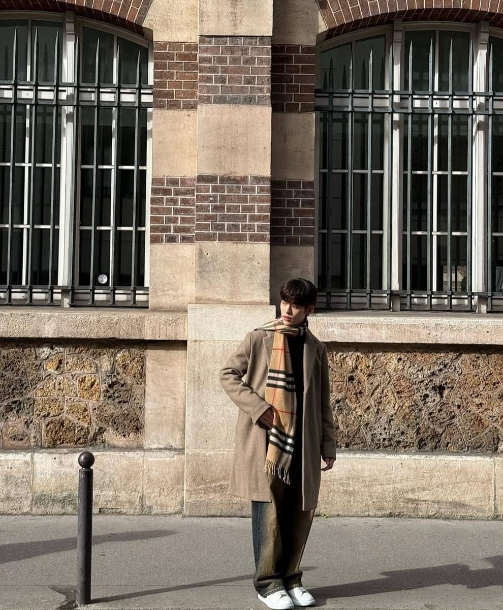 Seorang pria mengenakan mantel cokelat panjang, syal bermotif kotak, celana longgar, dan sepatu putih berdiri di depan bangunan bata.