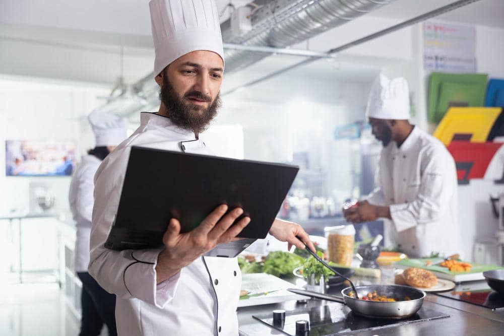 Seorang chef mengenakan seragam putih dan topi koki sedang memasak sambil memegang laptop di dapur profesional modern.