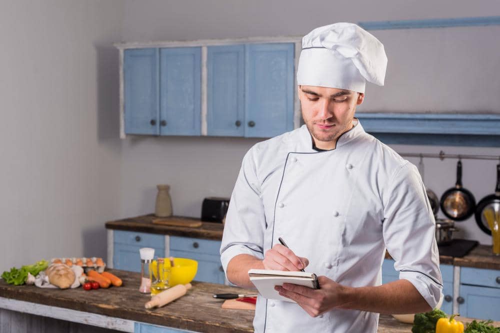 Seorang chef mengenakan seragam putih dan topi koki sedang menulis catatan di dapur dengan meja berisi bahan masakan.