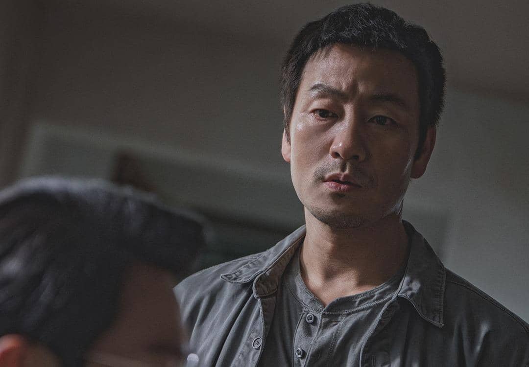 Seorang pria berwajah serius mengenakan kemeja abu-abu menatap seseorang di depannya dalam adegan drama Korea The Scarecrow.