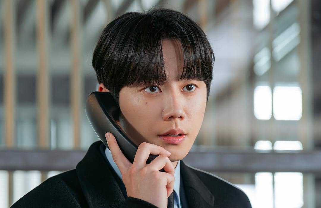 Seorang pria muda mengenakan setelan jas hitam sedang berbicara di telepon dengan ekspresi serius di dalam ruangan bercahaya alami.