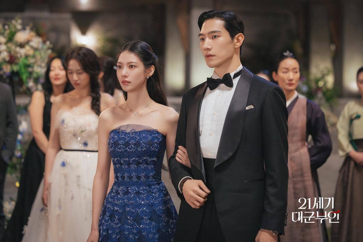 Dua pemeran drama Perfect Crown berdiri berdampingan di acara formal, mengenakan gaun biru elegan dan setelan jas hitam dengan dasi kupu-kupu.