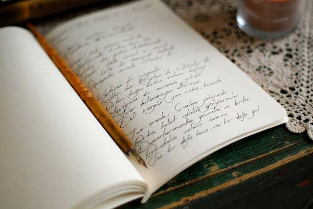 Buku catatan terbuka berisi tulisan tangan indah dengan pena kayu di atas meja kayu beralas taplak renda.
