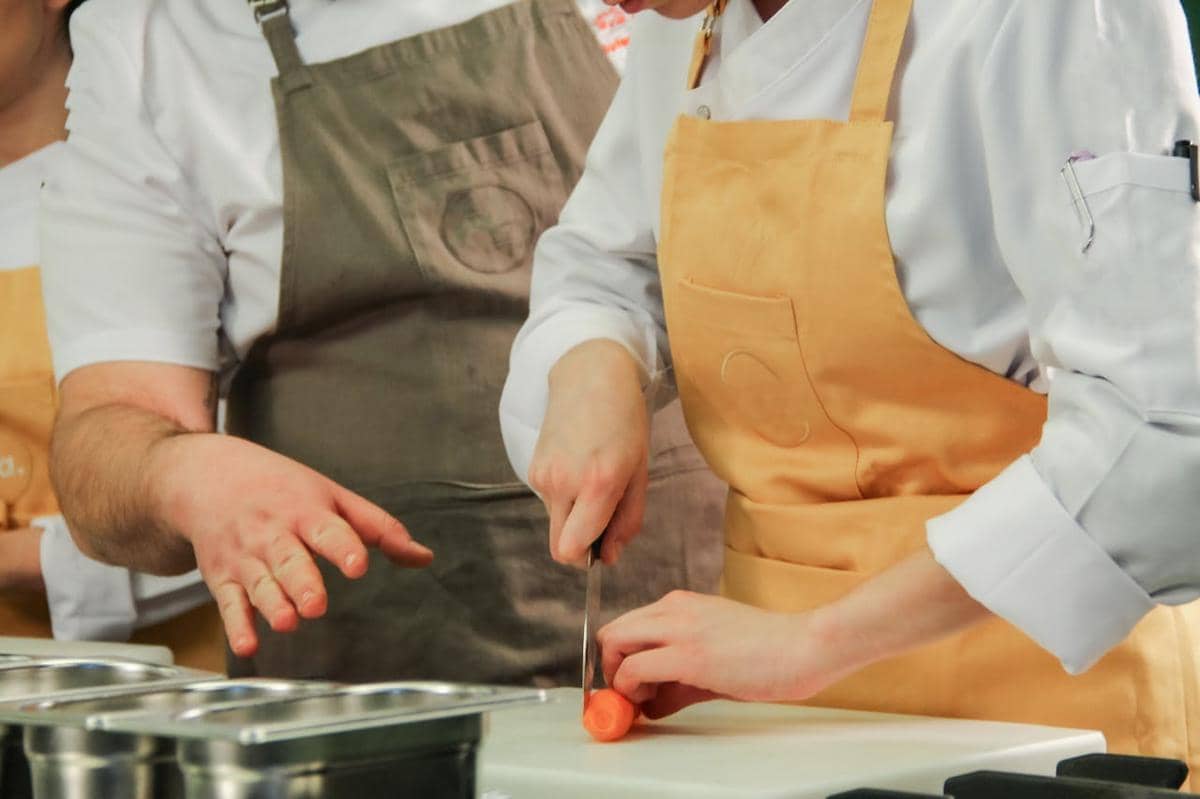 Seorang chef mengenakan celemek oranye sedang memotong wortel di dapur bersama rekan yang mengenakan celemek abu-abu.