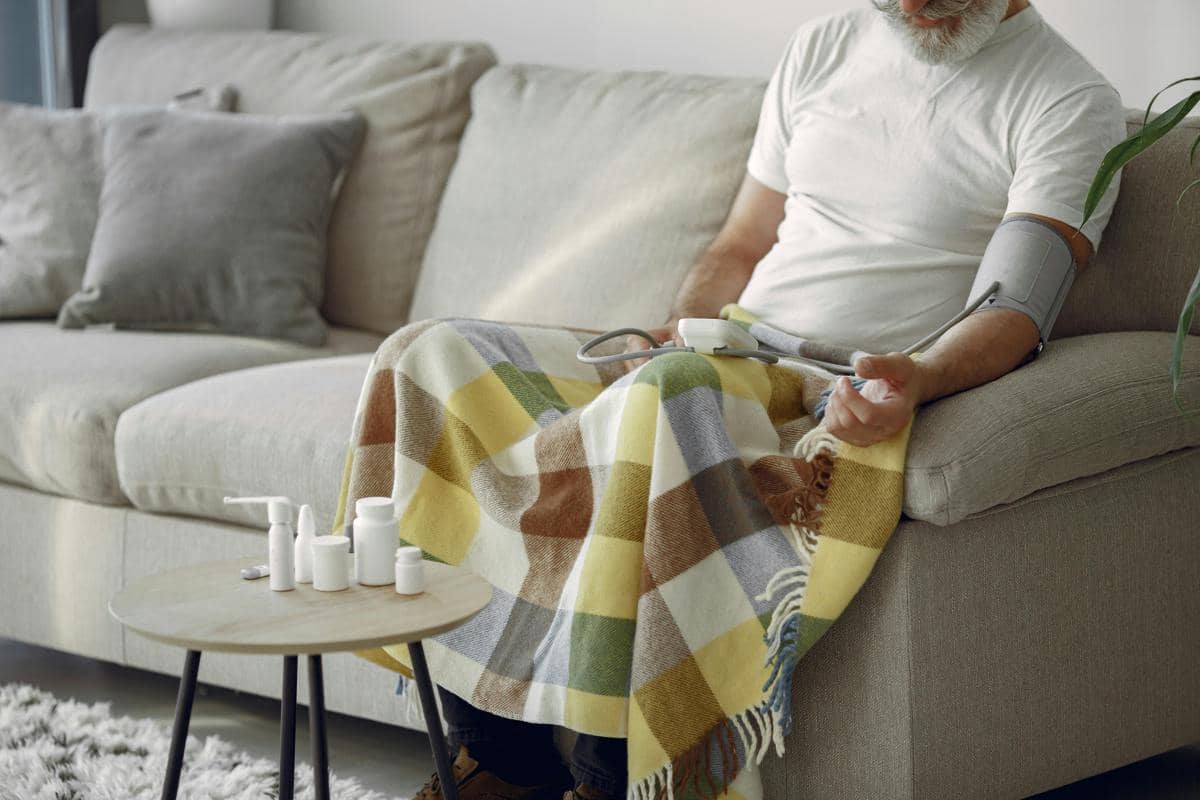 Seorang pria duduk di sofa sambil memeriksa tekanan darah menggunakan alat digital di rumah dengan selimut kotak warna-warni di pangkuannya.