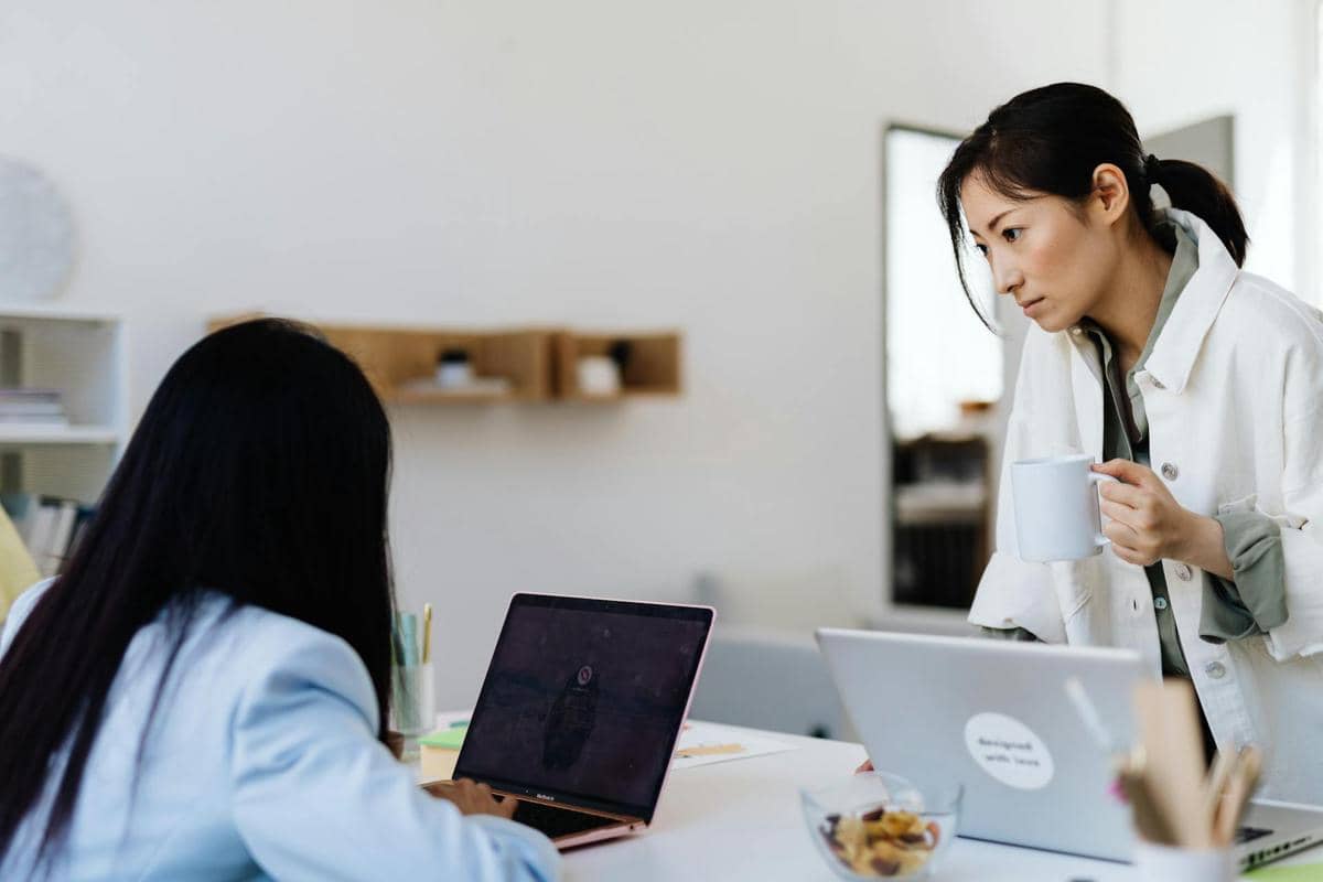 Seorang wanita berdiri sambil memegang cangkir dan menatap rekan kerjanya yang duduk di depan laptop di meja kantor modern.