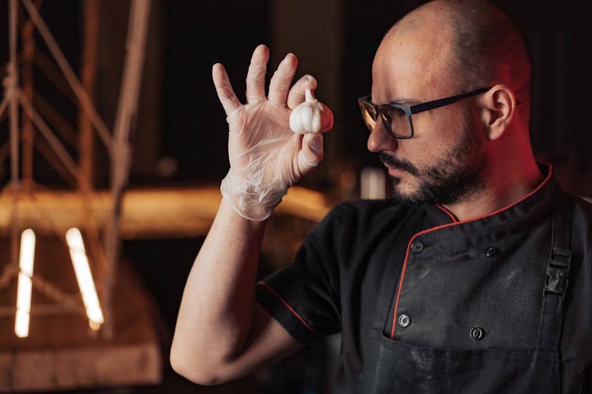 Seorang chef mengenakan seragam hitam dan sarung tangan lateks memegang bawang putih di dapur dengan pencahayaan hangat.