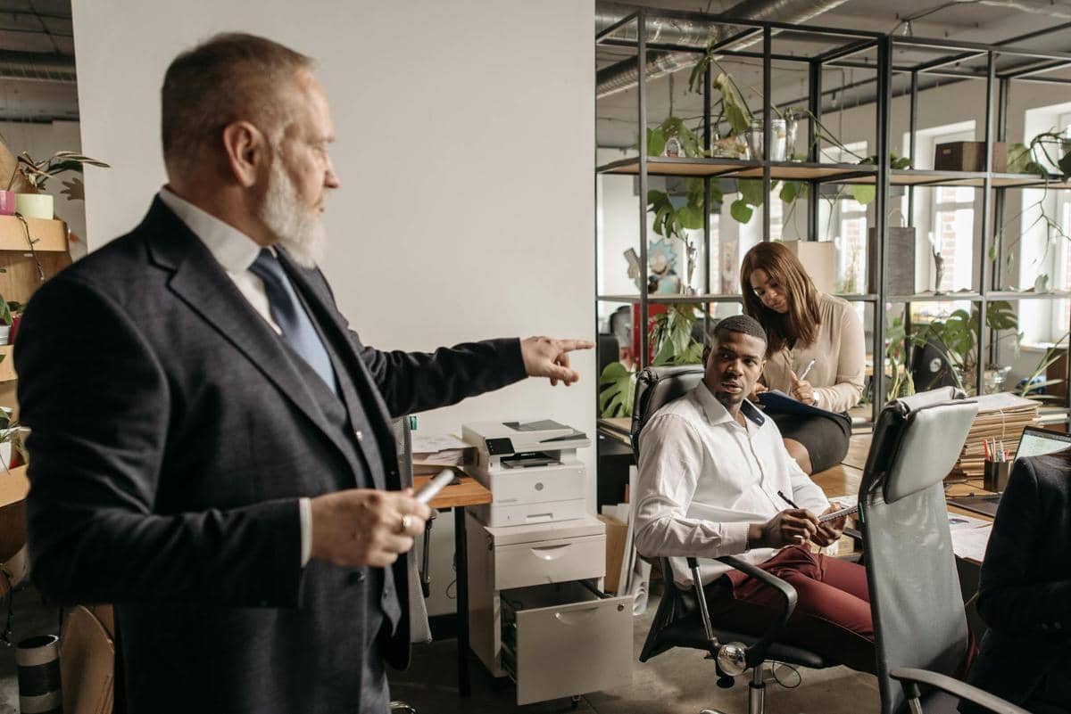 Seorang pria berjas menunjuk sambil berbicara kepada dua rekan kerja di kantor modern dengan suasana tegang dan serius.