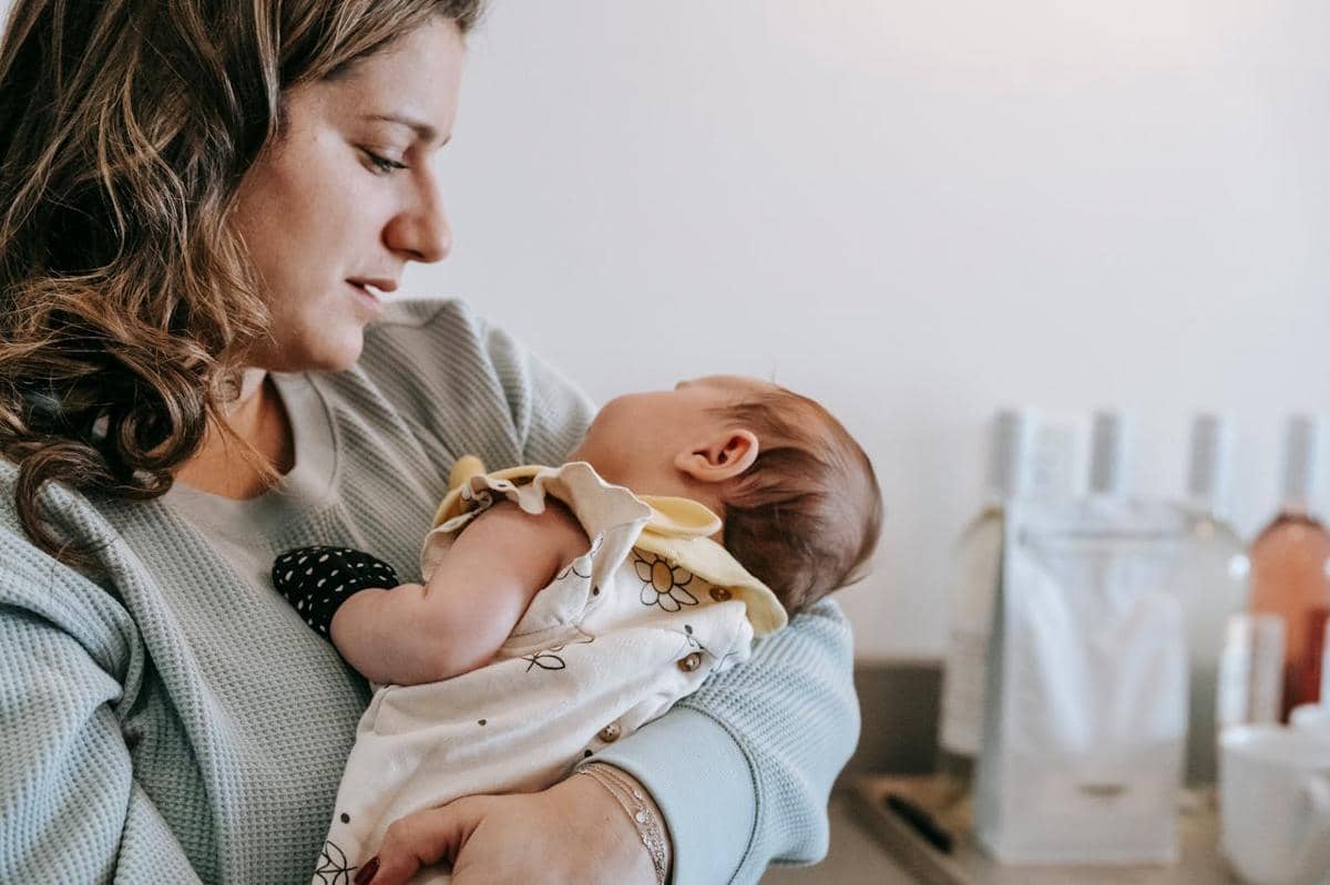 Seorang ibu memeluk bayinya dengan lembut di dalam ruangan, menatap wajah sang bayi yang tertidur dalam pelukannya.
