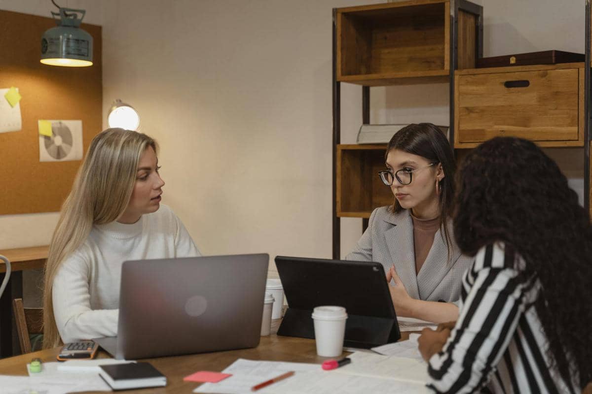 Tiga rekan kerja wanita berdiskusi di meja kantor dengan laptop, dokumen, dan cangkir kopi di ruangan kerja modern.