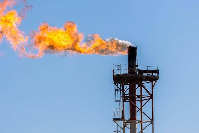 Api besar menyala dari pipa industri tinggi di bawah langit biru, menggambarkan pembakaran gas metana di fasilitas energi.