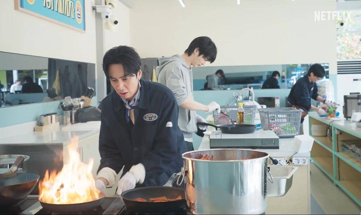 Beberapa peserta variety show sedang memasak di dapur modern, salah satunya menggoreng dengan api besar di atas kompor.