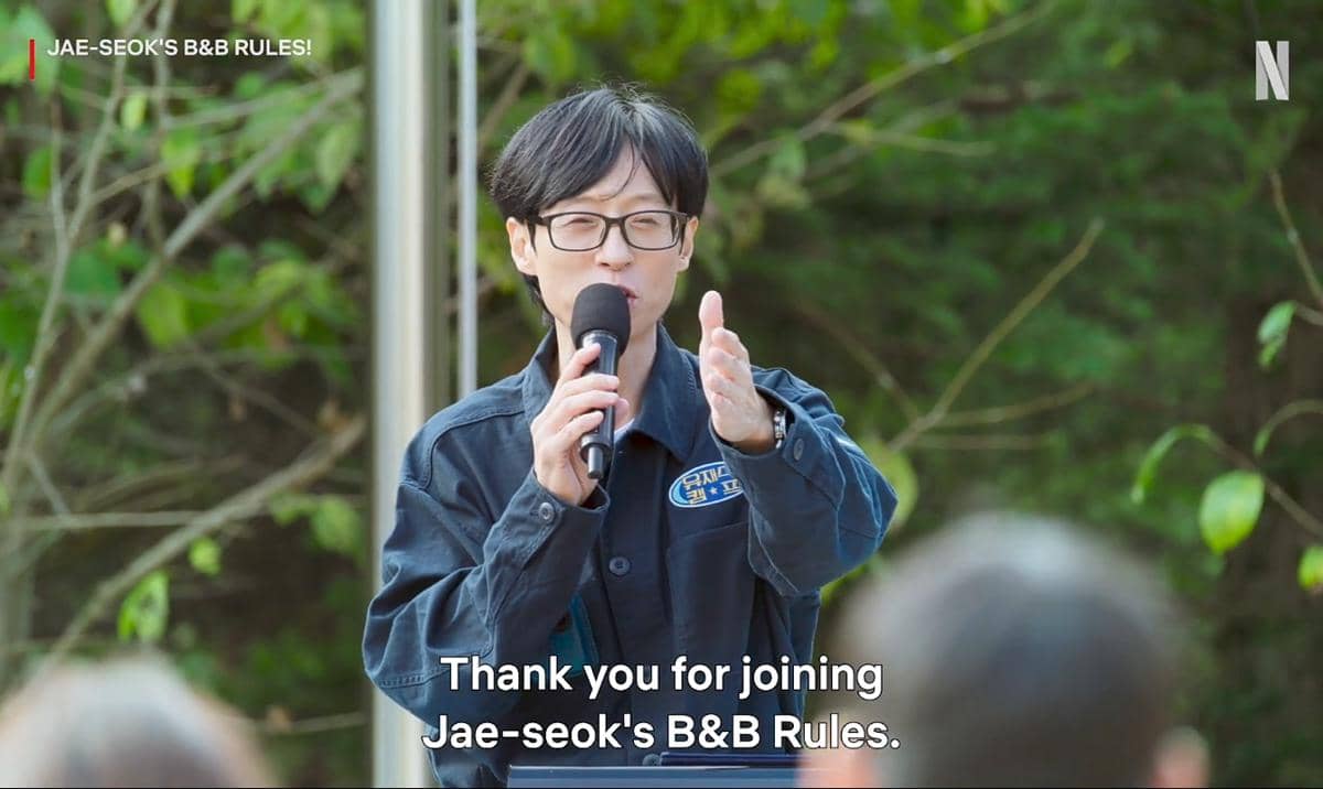 Seorang pembawa acara mengenakan jaket biru tua dan kacamata sedang berbicara dengan mikrofon di luar ruangan berlatar pepohonan.