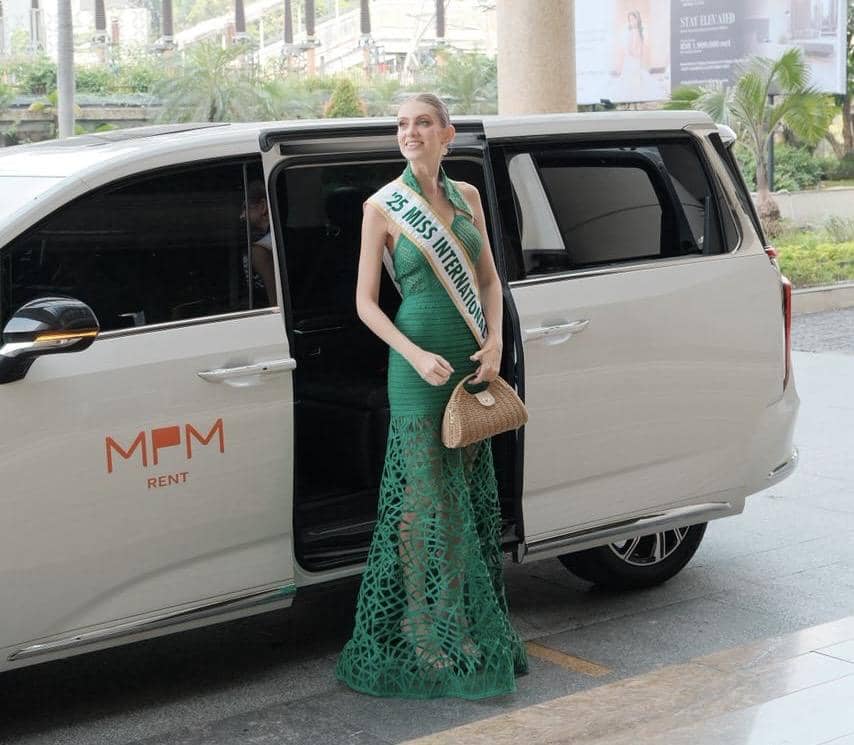 Seorang finalis Putri Indonesia mengenakan gaun hijau berdiri di samping mobil MPMRent berwarna putih dengan pintu terbuka.