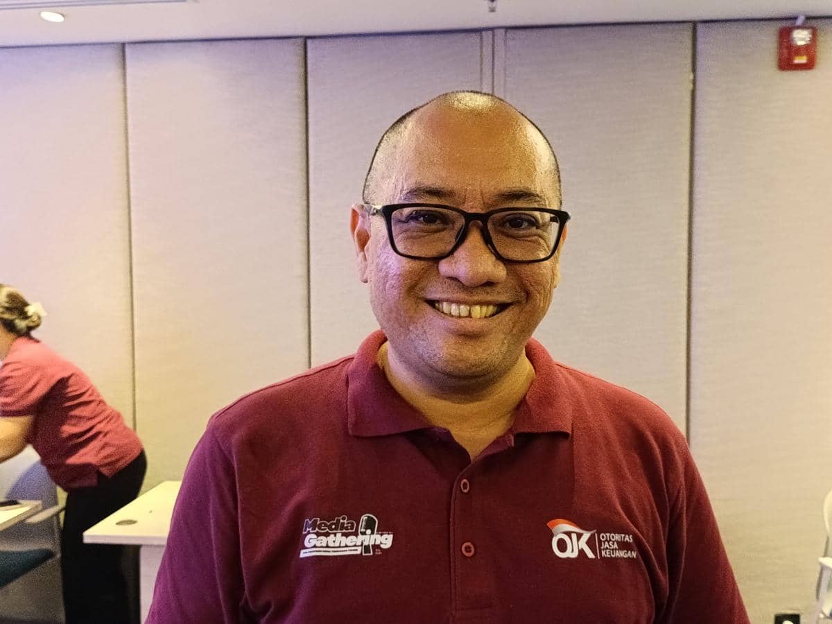 Seorang pria mengenakan kacamata dan kaus polo merah berlogo Otoritas Jasa Keuangan sedang tersenyum di dalam ruangan.