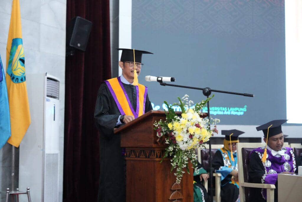 Seorang dosen Universitas Lampung mengenakan toga sedang menyampaikan pidato di podium dalam acara pengukuhan guru besar di GSG Unila.