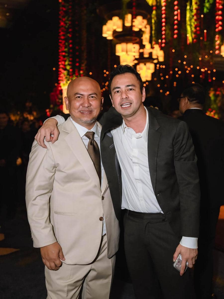 Dua pria mengenakan setelan jas berpose bersama di acara malam dengan pencahayaan hangat dan dekorasi lampu gantung.