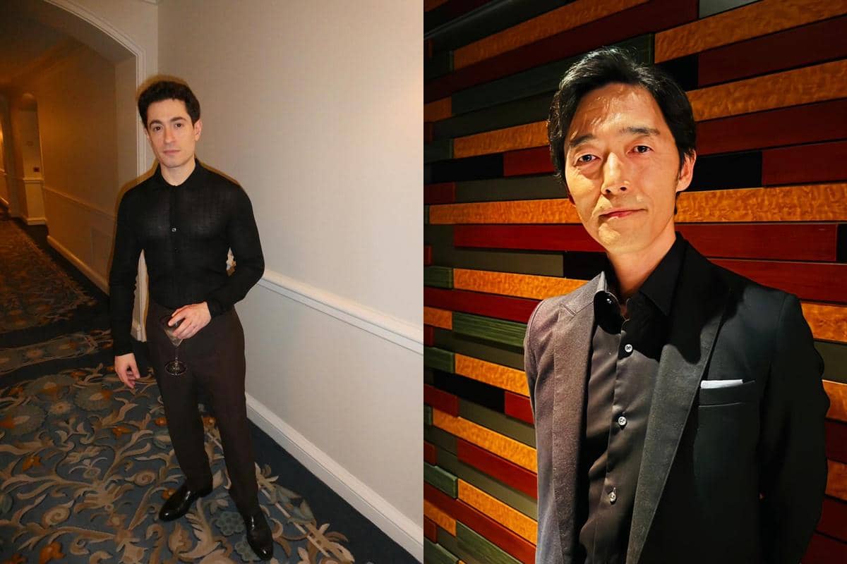 Dua pria berpose terpisah, satu berdiri di lorong hotel dengan pakaian hitam dan memegang gelas, satunya di depan dinding berwarna kayu.