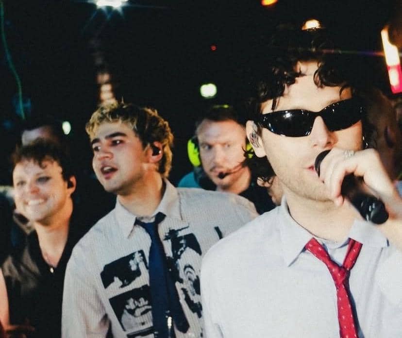 Beberapa anggota band tampil di atas panggung dengan pencahayaan warna-warni, salah satu mengenakan kacamata hitam dan memegang mikrofon.