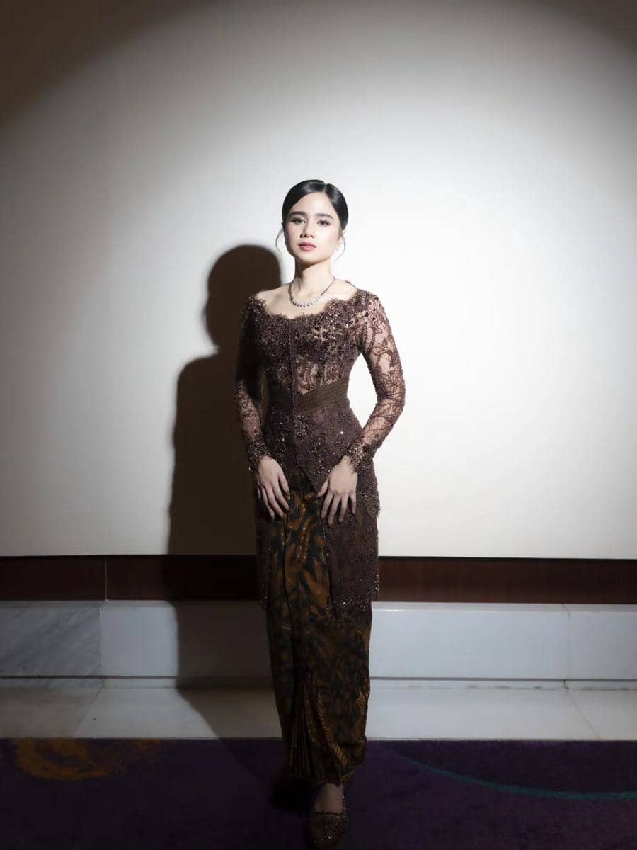 Seorang perempuan mengenakan kebaya cokelat berhias payet dan kain batik berdiri di ruangan dengan pencahayaan lembut.