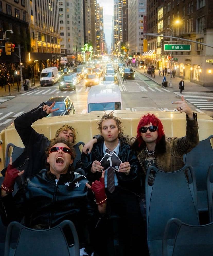 Empat pria bergaya rock duduk di bus tingkat terbuka di tengah kota besar dengan latar gedung tinggi dan lalu lintas padat.