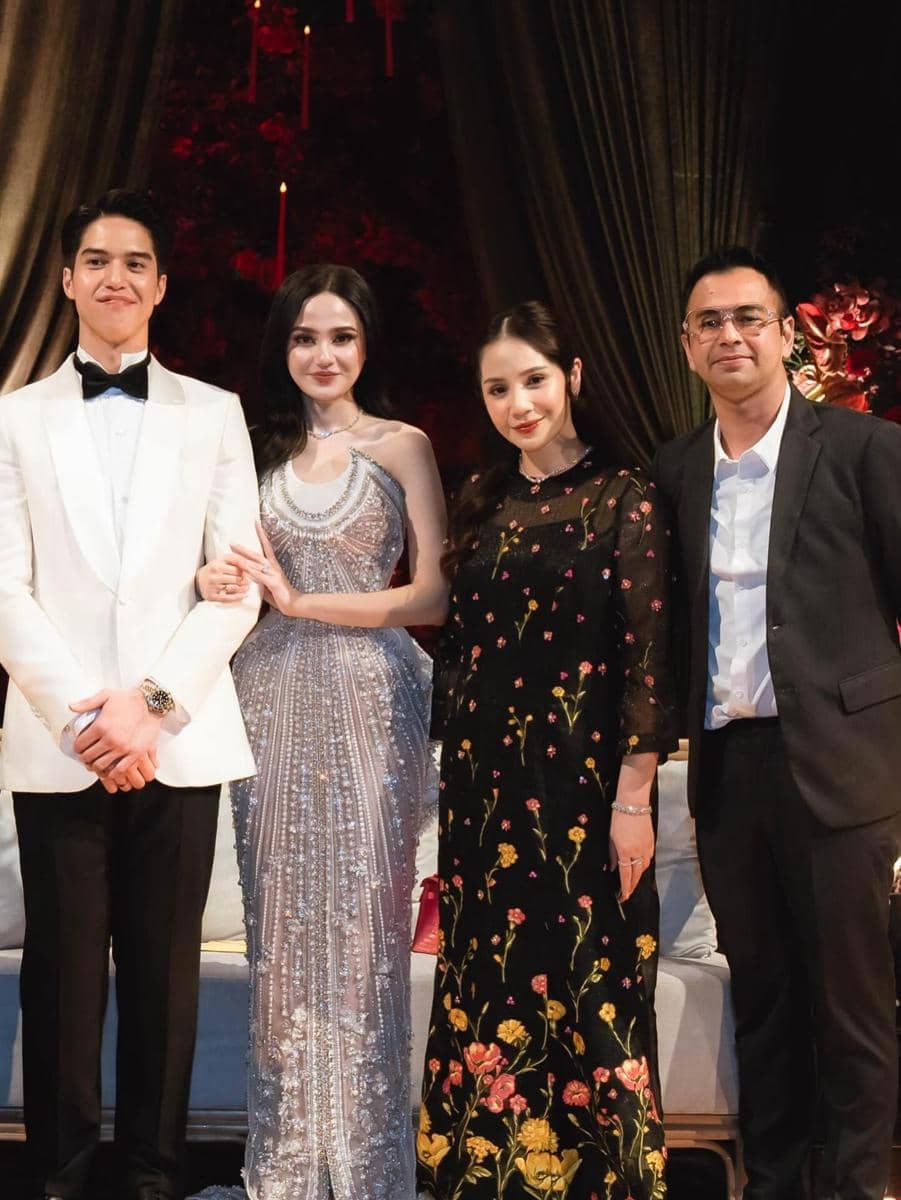 Empat orang berpose bersama di acara pernikahan, dua pria mengenakan jas formal dan dua wanita memakai gaun elegan di latar dekorasi mewah.