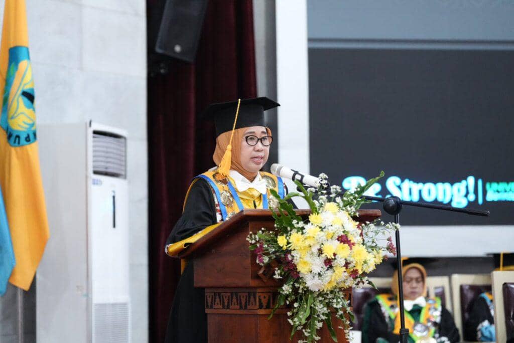 Seorang dosen Unila mengenakan toga akademik menyampaikan pidato di podium berhias bunga pada acara pengukuhan guru besar di GSG Unila.