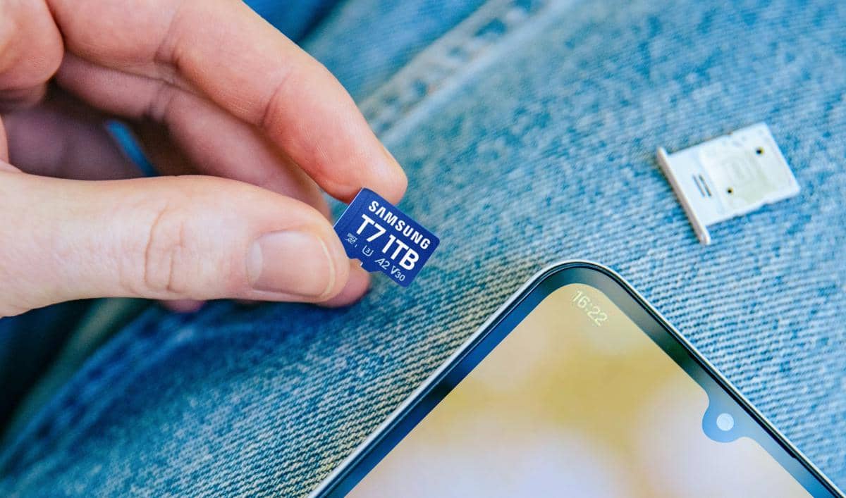 Seseorang memegang kartu memori Samsung MicroSD T7 berkapasitas 1TB di atas permukaan kain denim dengan ponsel dan slot kartu di dekatnya.