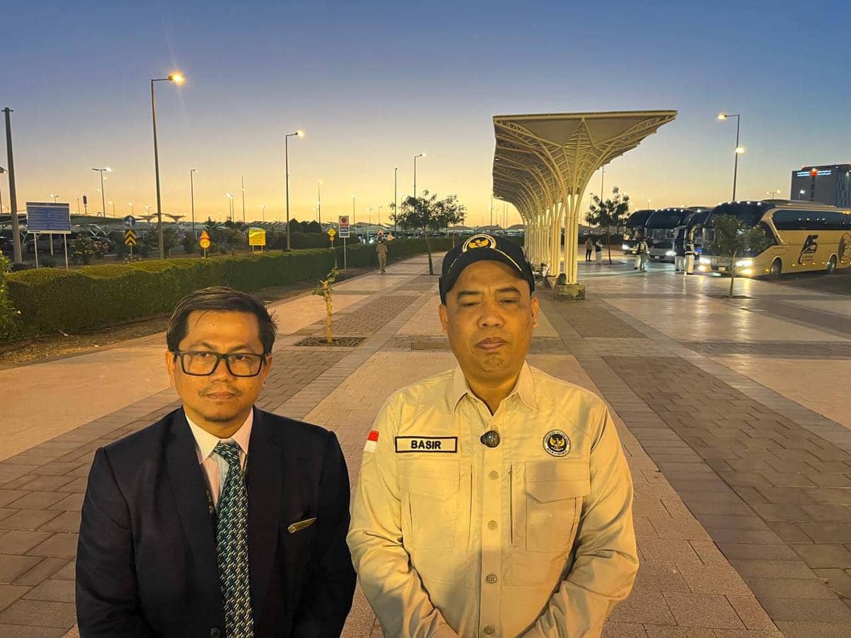 Dua pria berdiri di area luar bandara saat senja, salah satunya mengenakan jas dan dasi, sementara yang lain memakai seragam krem dan topi hitam.