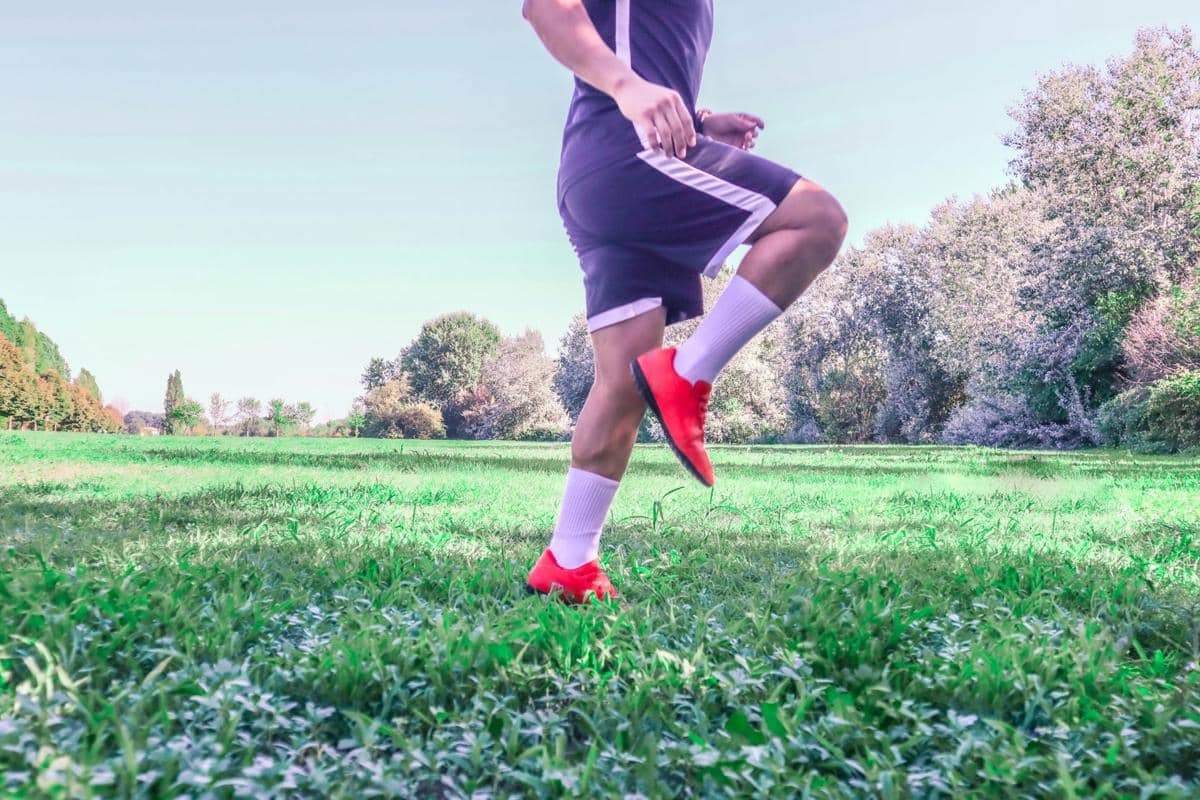 Seorang pria berolahraga melakukan latihan lari di lapangan rumput hijau dengan sepatu merah dan kaus kaki putih.