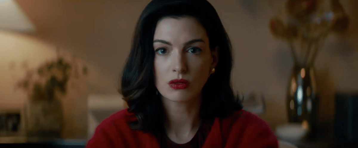 Seorang wanita berambut hitam dengan riasan tegas dan mengenakan pakaian merah duduk di ruangan dengan pencahayaan hangat.