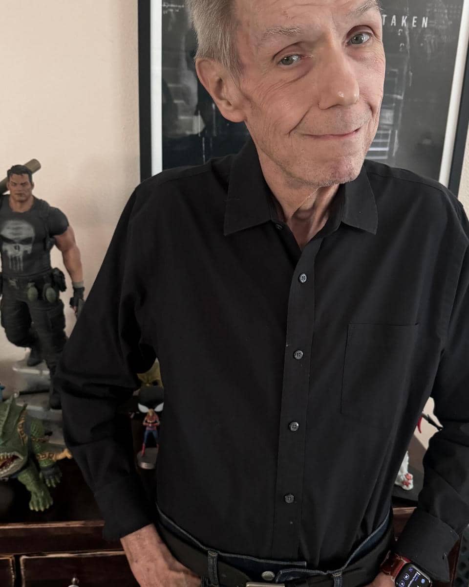 Seorang pria berambut abu-abu mengenakan kemeja hitam berdiri di dalam ruangan dengan beberapa figur aksi di meja di belakangnya.