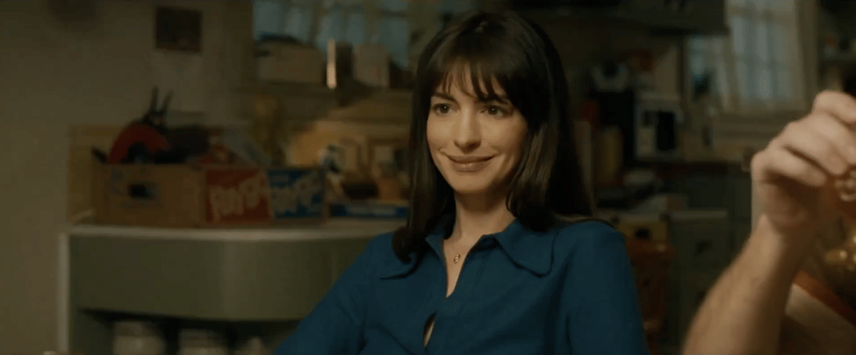 Seorang wanita berambut hitam tersenyum sambil duduk di meja makan dalam adegan film The End of Oak Street.