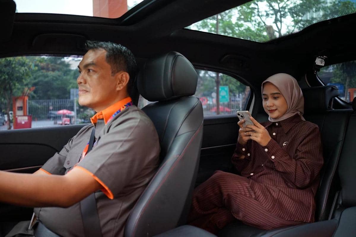 Seorang pengemudi membawa penumpang wanita berhijab yang sedang menggunakan ponsel di dalam mobil dengan konsep ramah lingkungan.